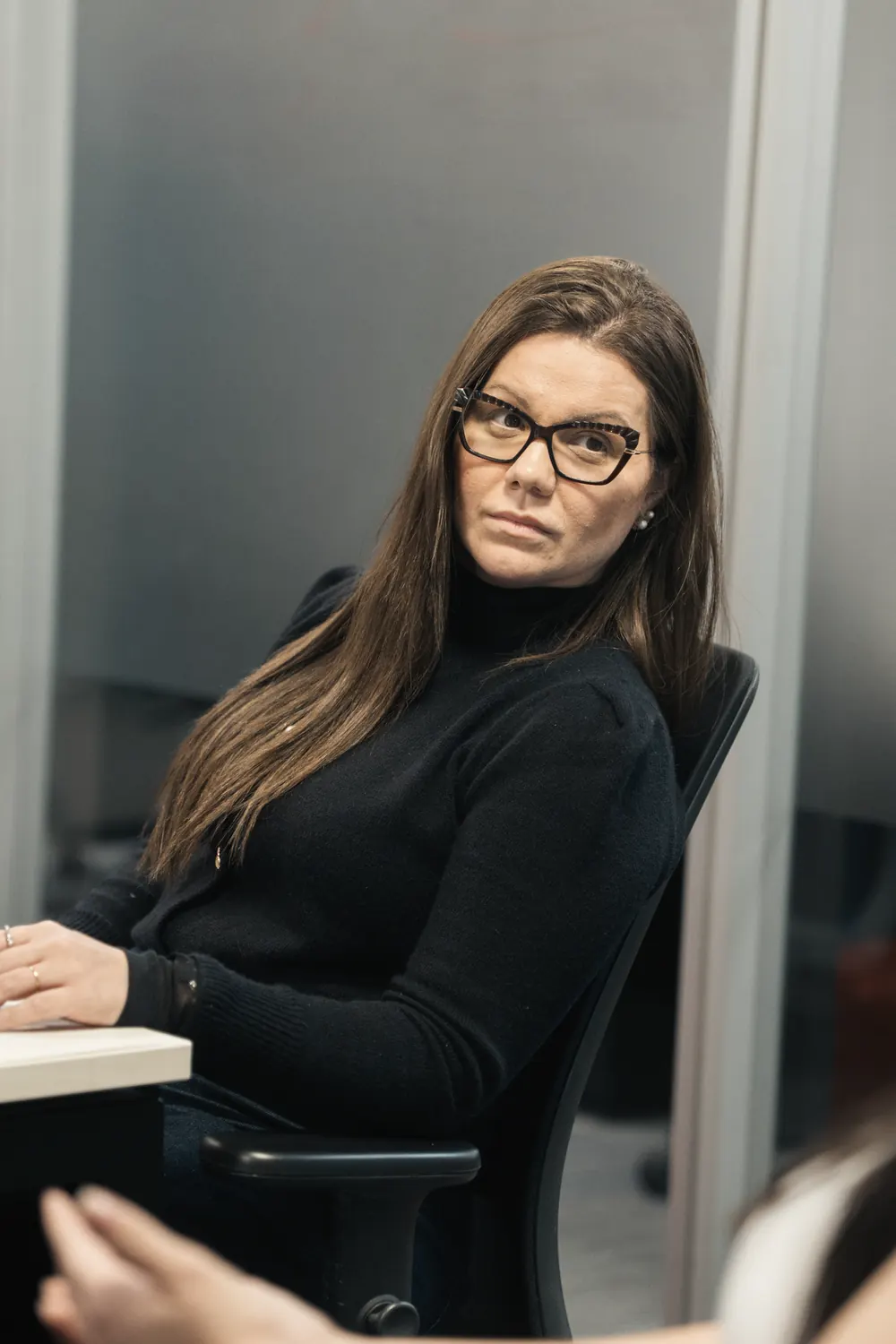 Nathália Gomes sentada à mesa de trabalho, de blusa preta e óculos, olhando para o lado.