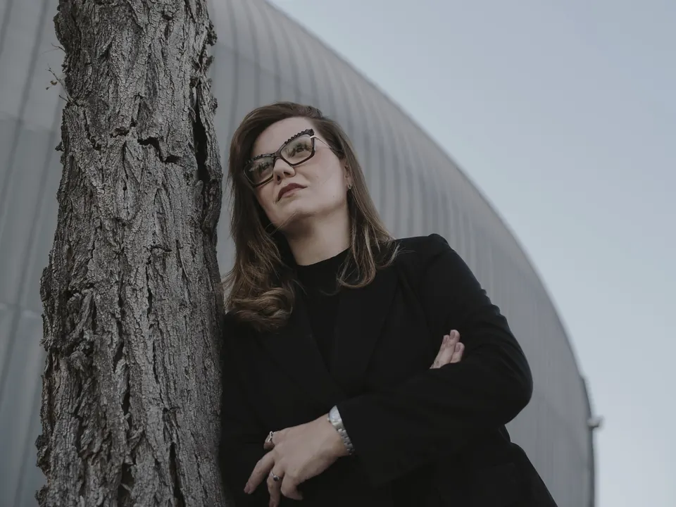 Nathália Gomes ao ar livre, de braços cruzados, ao lado de uma árvore.