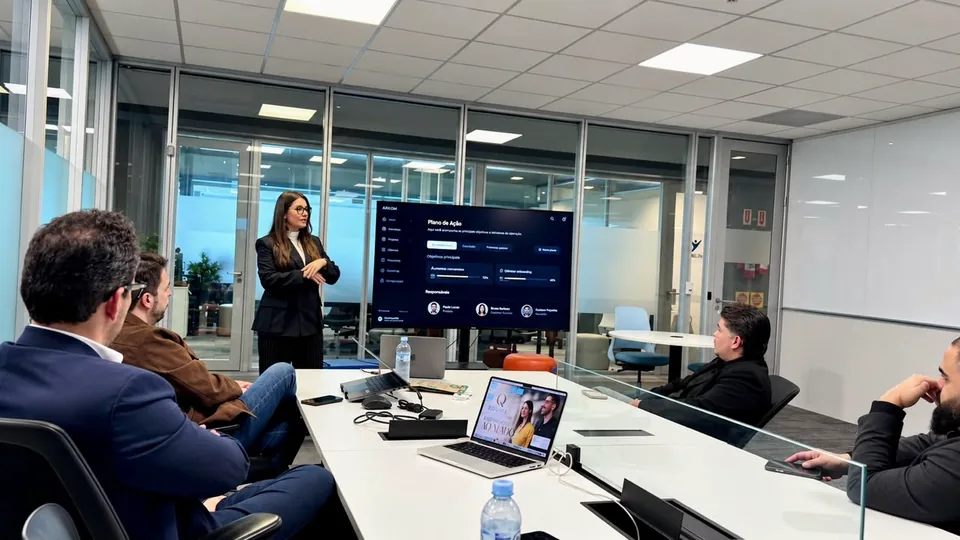 Nathália Gomes de pé, conduzindo uma reunião com o time em volta da mesa, com uma tela ao fundo.